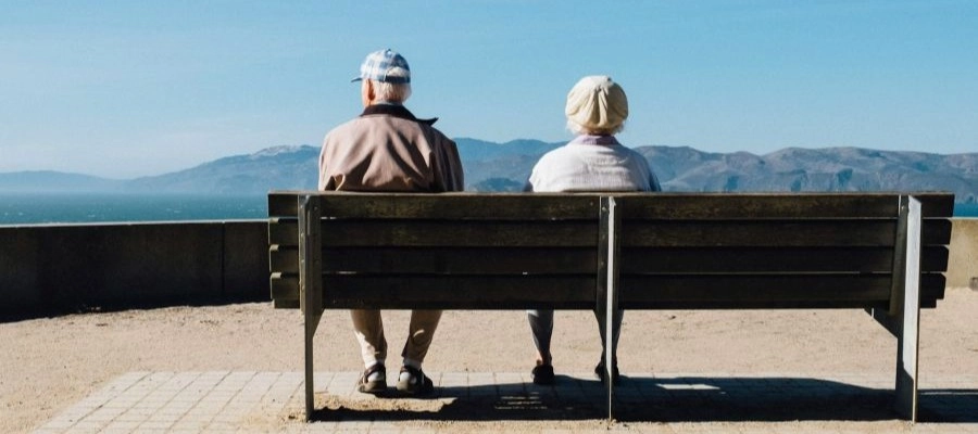 Na imagem, um senhor e uma senhora estão sentados em um banco de frente para o mar.