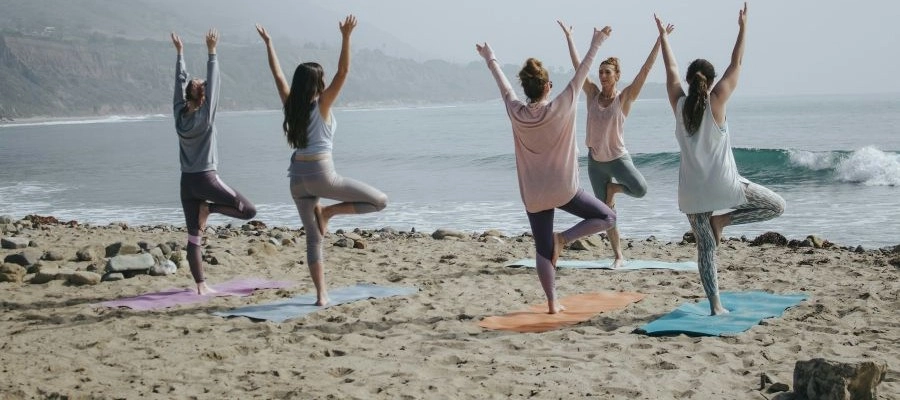Pessoas praticando yoga na praia, em frente ao mar, com posturas de equilíbrio e braços erguidos, representando bem-estar, wellness, energia e conexão com a natureza.